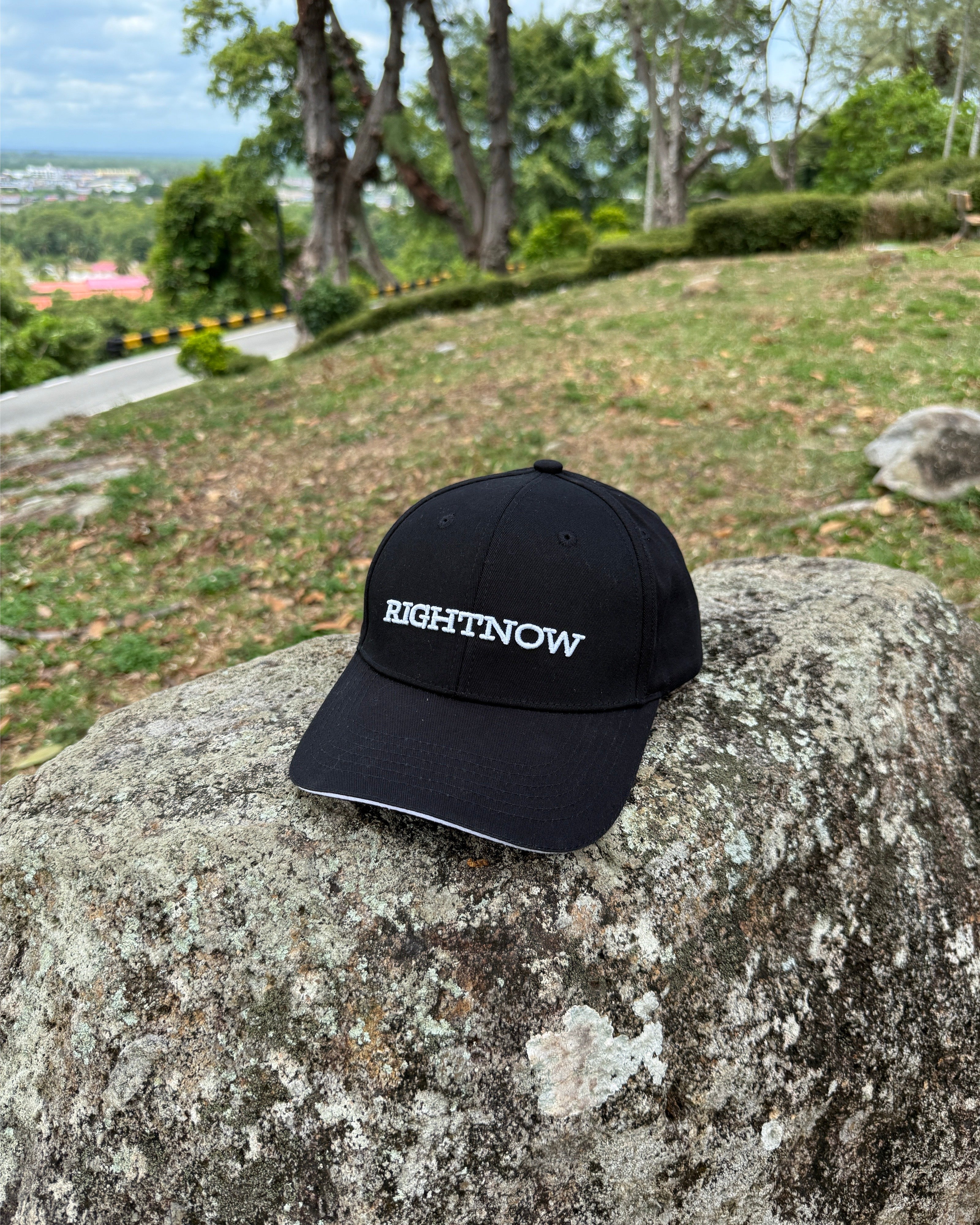 A matt black Ignite RIGHTNOW logo cap on a mountain rock at a scenic lookout point, a simple accessory for being present and enjoying an authentic view.