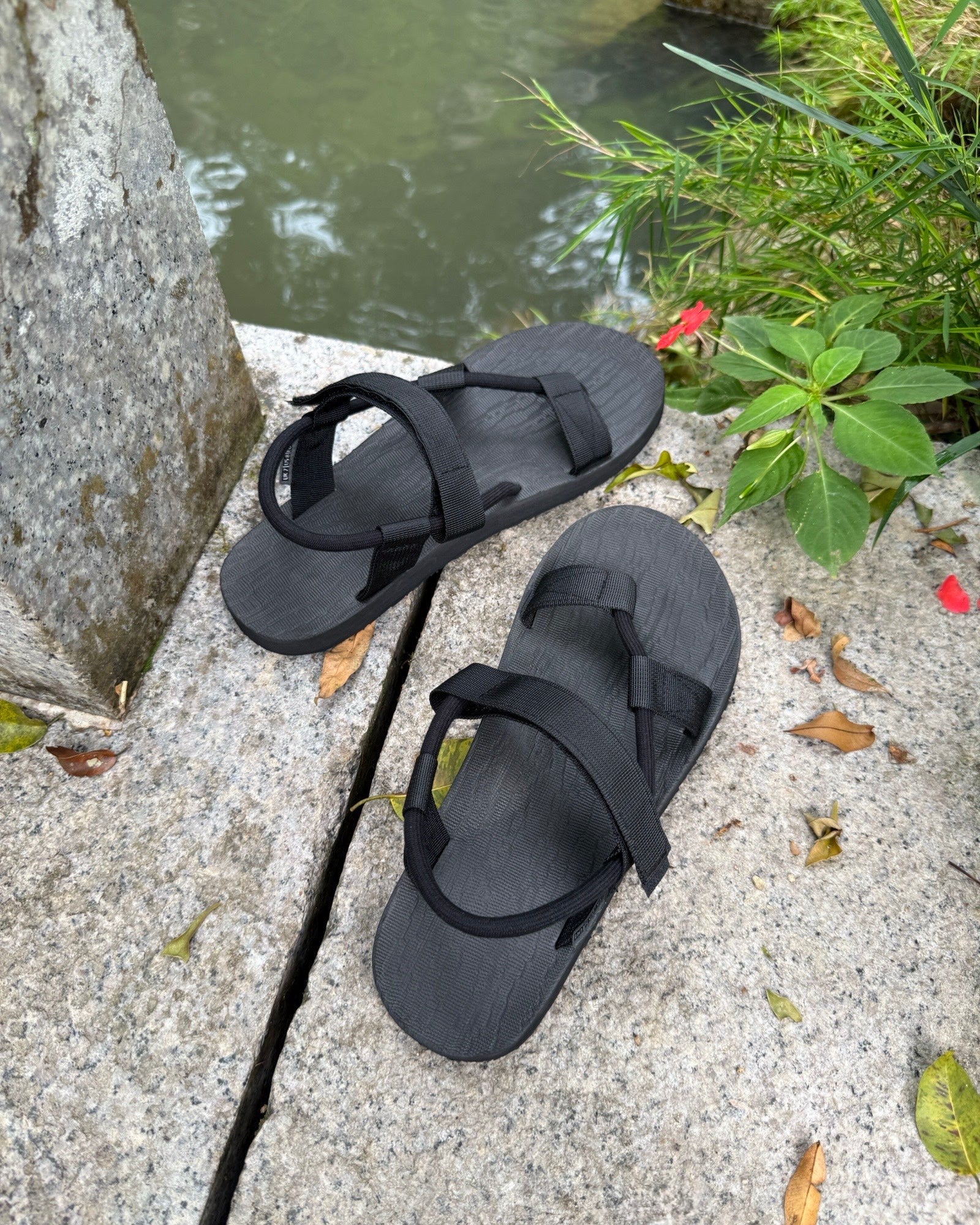 A pair of men's matt black EXPLOre Roman Sandals on a stone ledge by a tranquil stream, showing a simple, authentic lifestyle connected with nature.