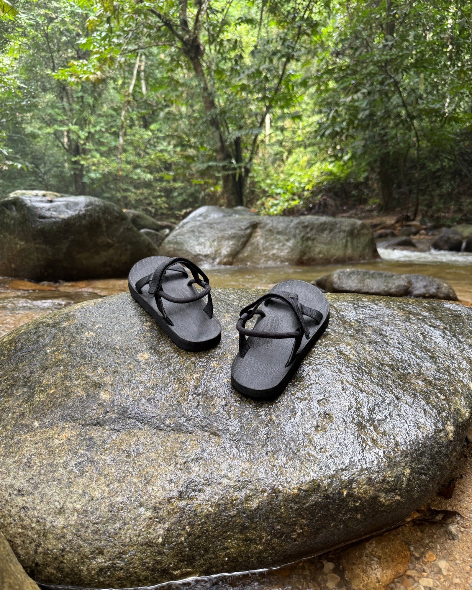 Men's matt black EXPLOre Roman Sandals on a river rock with a lush tropical rainforest backdrop, the perfect footwear for authentic tropical exploration.