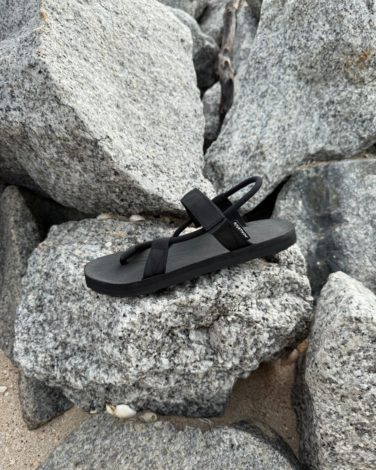 A single men's matt black EXPLOre Roman Sandal on a rugged coastal granite boulder, highlighting its durable minimalist strap design for all-terrain wear.