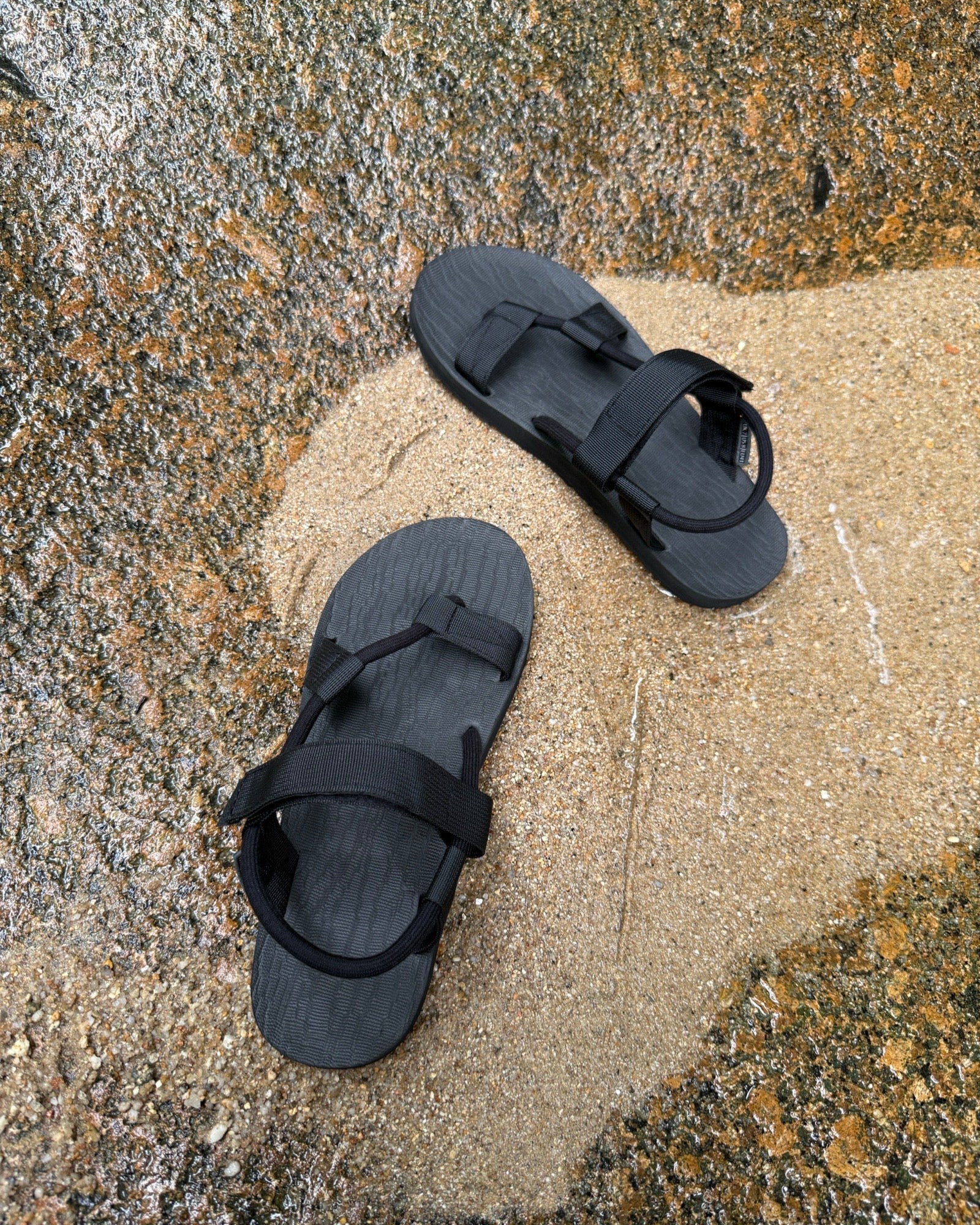 Top-down view of men's elegant-tone EXPLOre Roman Sandals on wet, sandy riverside ground, showing their simple design and flexible straps for any natural terrain.