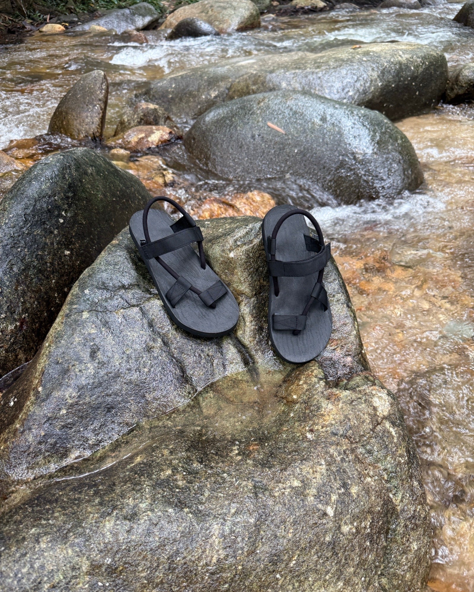 Waterproof men's matt black EXPLOre Roman Sandals on a wet boulder in a flowing jungle stream, highlighting their anti-slip soles for secure footing on any terrain.