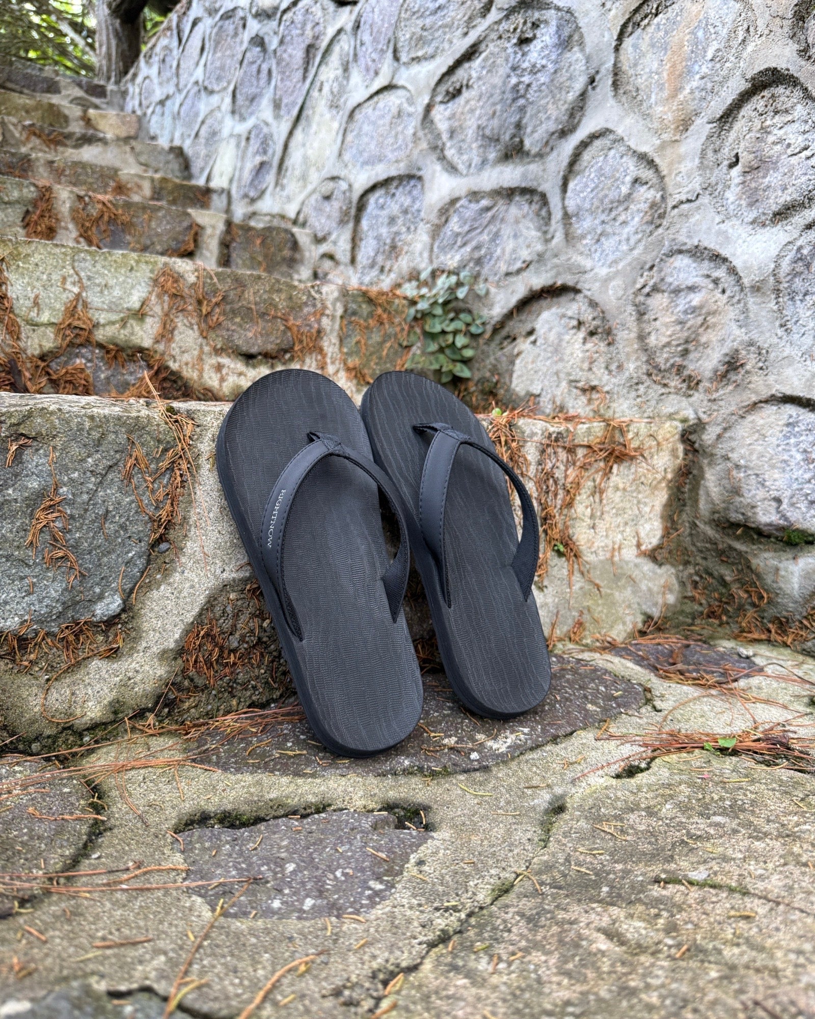 A pair of men's matt black Flow Thong Sandals on rustic stone steps, perfect for a simple and authentic journey through historical or natural paths.