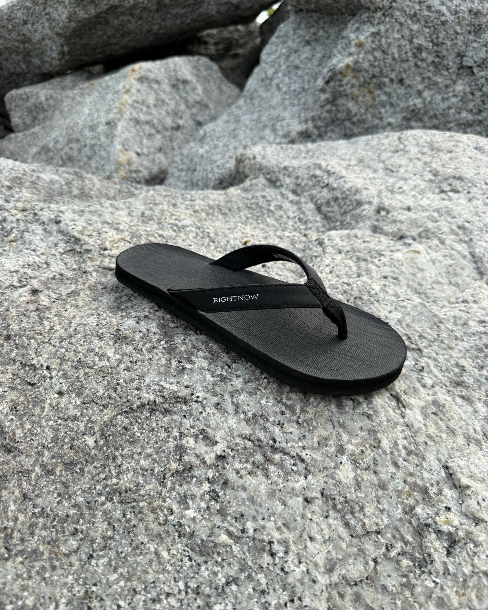 A single men's matt black Flow Thong Sandal resting on rugged coastal rock, showcasing its minimalist design for effortless outdoor presence.