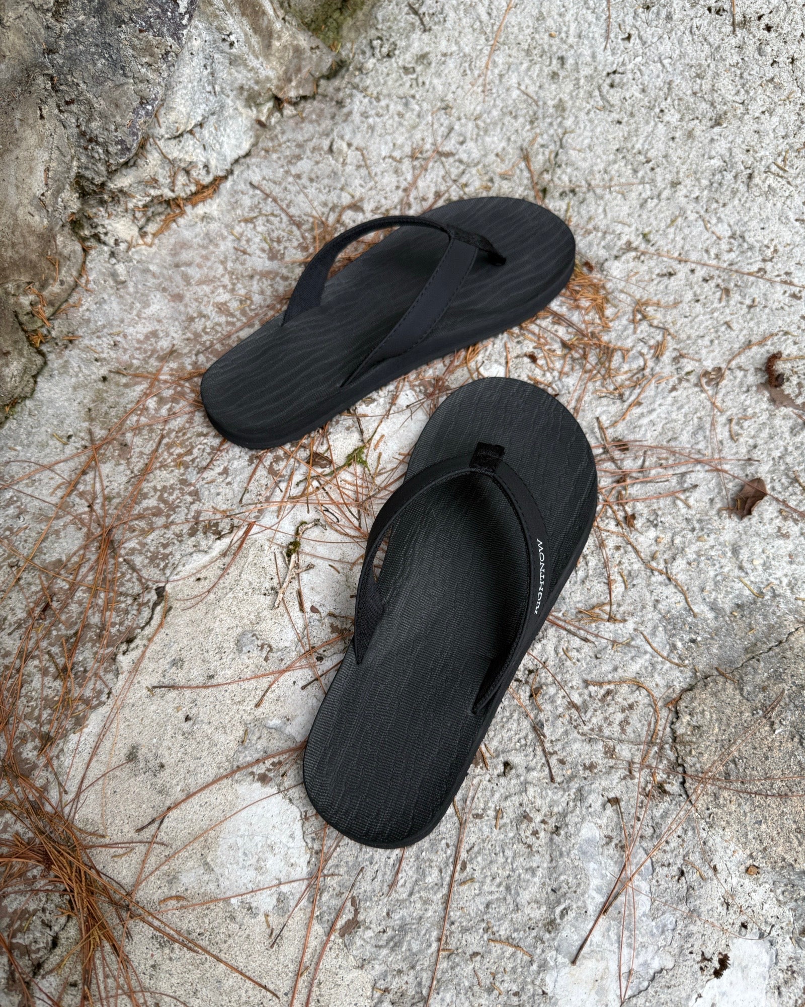 Top-down view of men's elegant-tone Flow Thong Sandals on concrete ground with scattered pine needles, highlighting their simple design for natural and urban exploration.