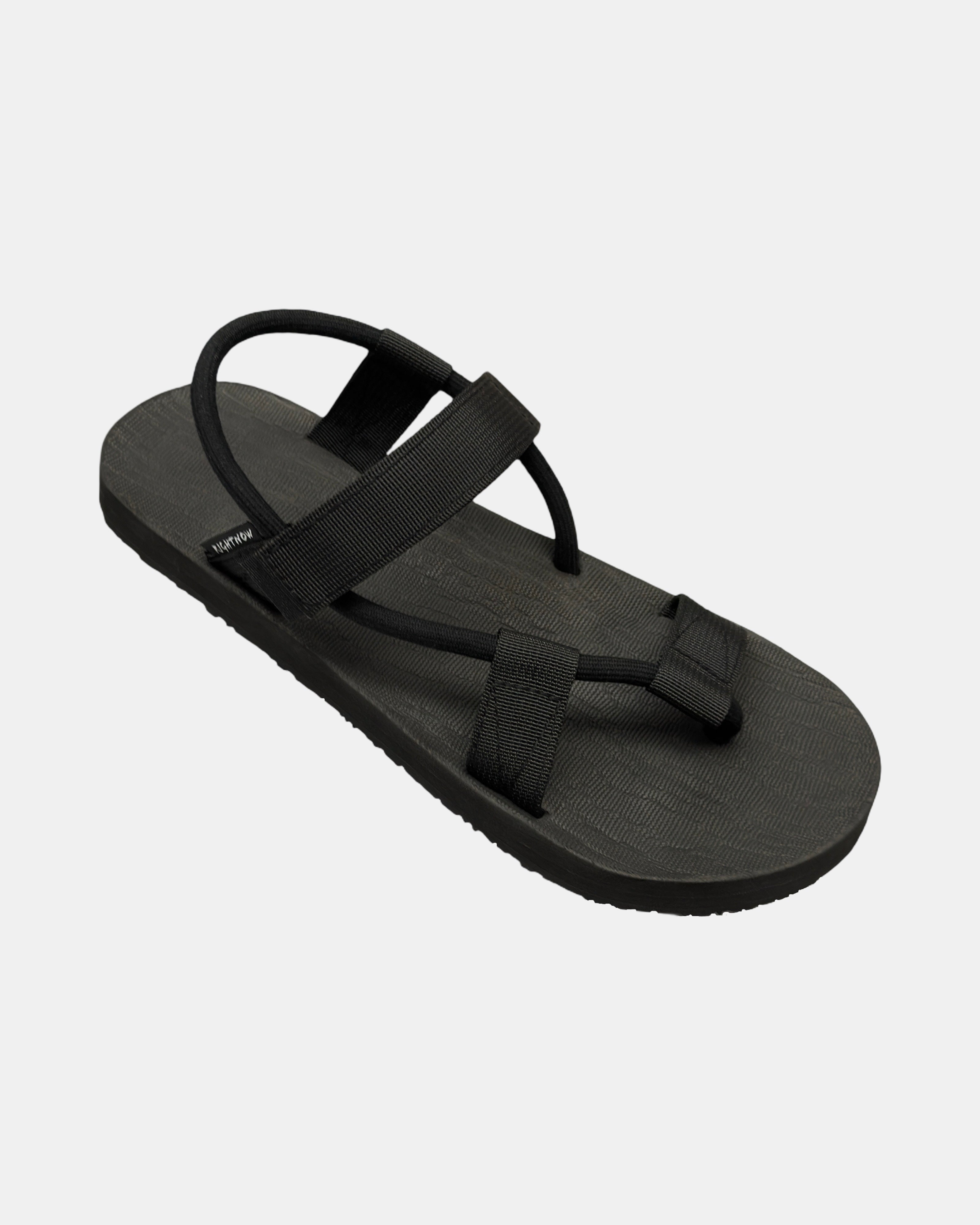 A 45-degree angled view of a men's matt black rubber Roman-style sandal with a single 360° flexible strap around the ankle.