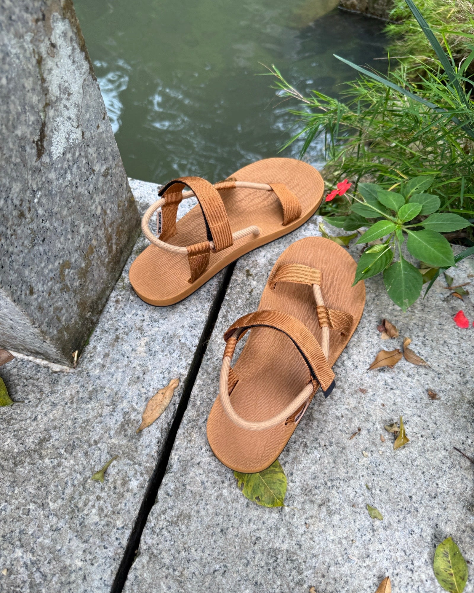 A pair of men's earth brown EXPLOre Roman Sandals on a stone ledge by a tranquil stream, showing a simple, authentic lifestyle connected with nature.