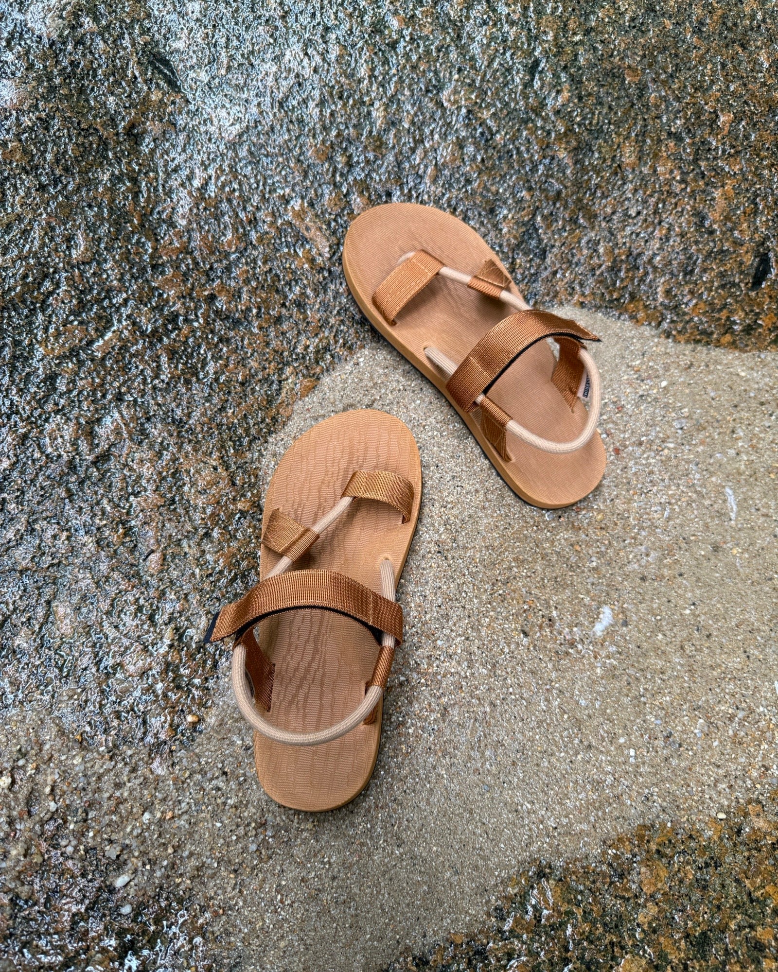 Top-down view of men's earth-tone EXPLOre Roman Sandals on wet, sandy riverside ground, showing their simple design and flexible straps for any natural terrain.