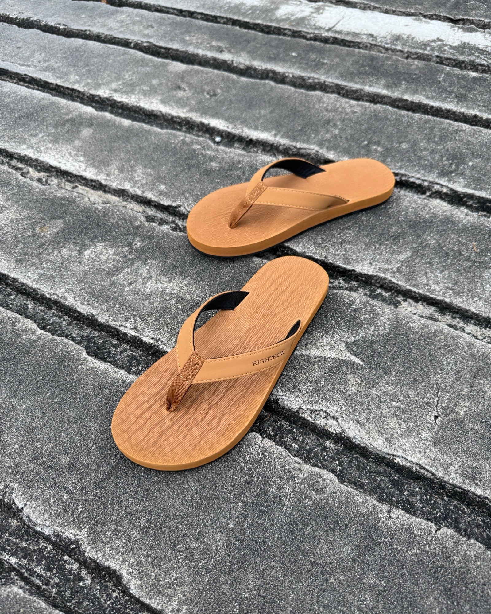 A pair of men's earth brown Flow Thong Sandals on a dark, textured concrete surface, showcasing their minimalist comfort for modern living.