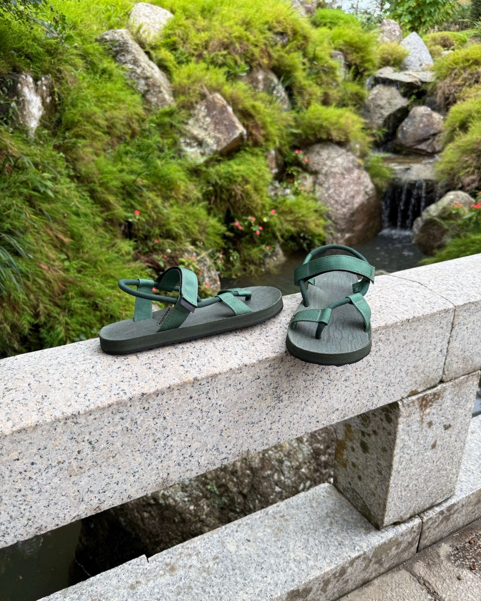 Men's natural green EXPLOre Roman Sandals on a bridge over a serene waterfall and lush greenery, perfect for travel and finding a quiet moment of presence in nature.