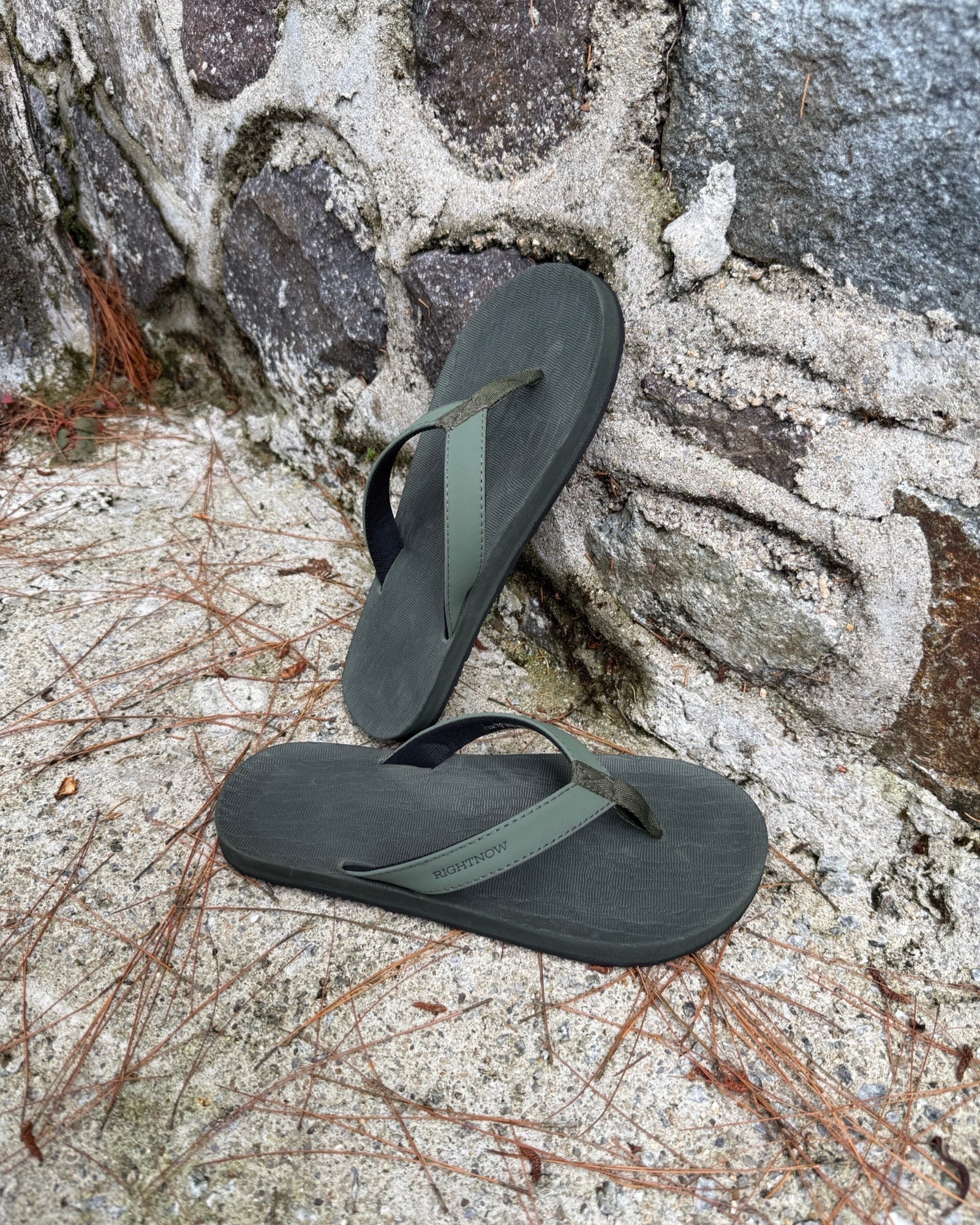 Men's natural green Flow Thong Sandals leaning against a textured stone wall, conveying a casual yet authentic style ready for everyday adventures.