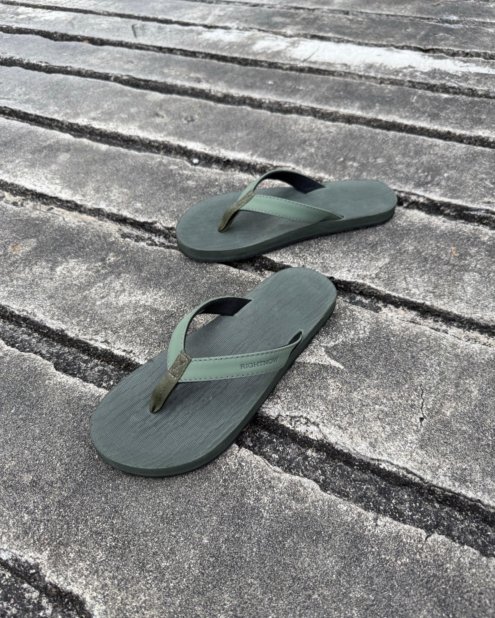 A pair of men's natural green Flow Thong Sandals on a dark, textured concrete surface, showcasing their minimalist comfort for modern living.