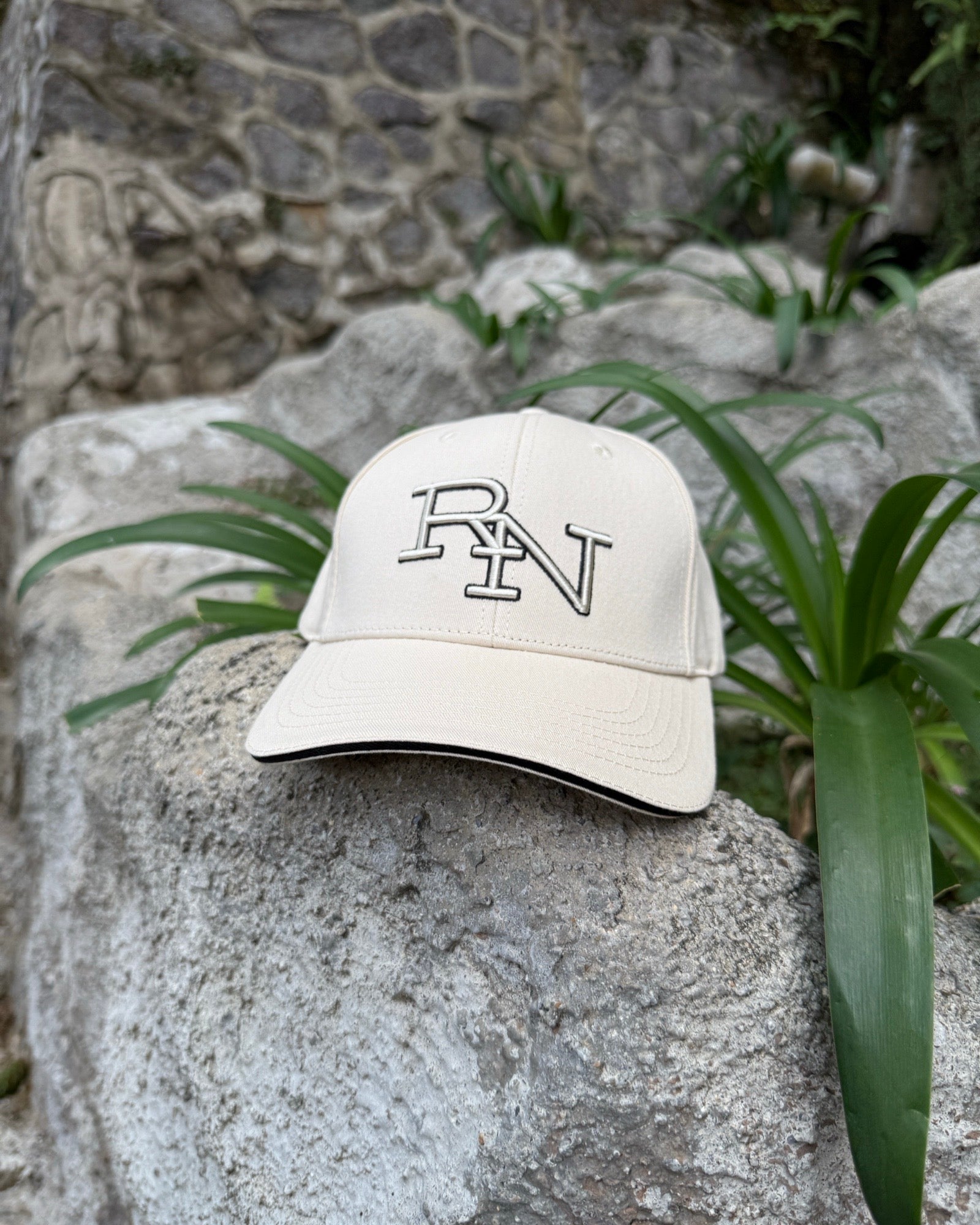 A sand beige Ignite RN logo cap resting on a large, sunlit rock in a quiet garden, embodying a moment of simple, peaceful presence outdoors.