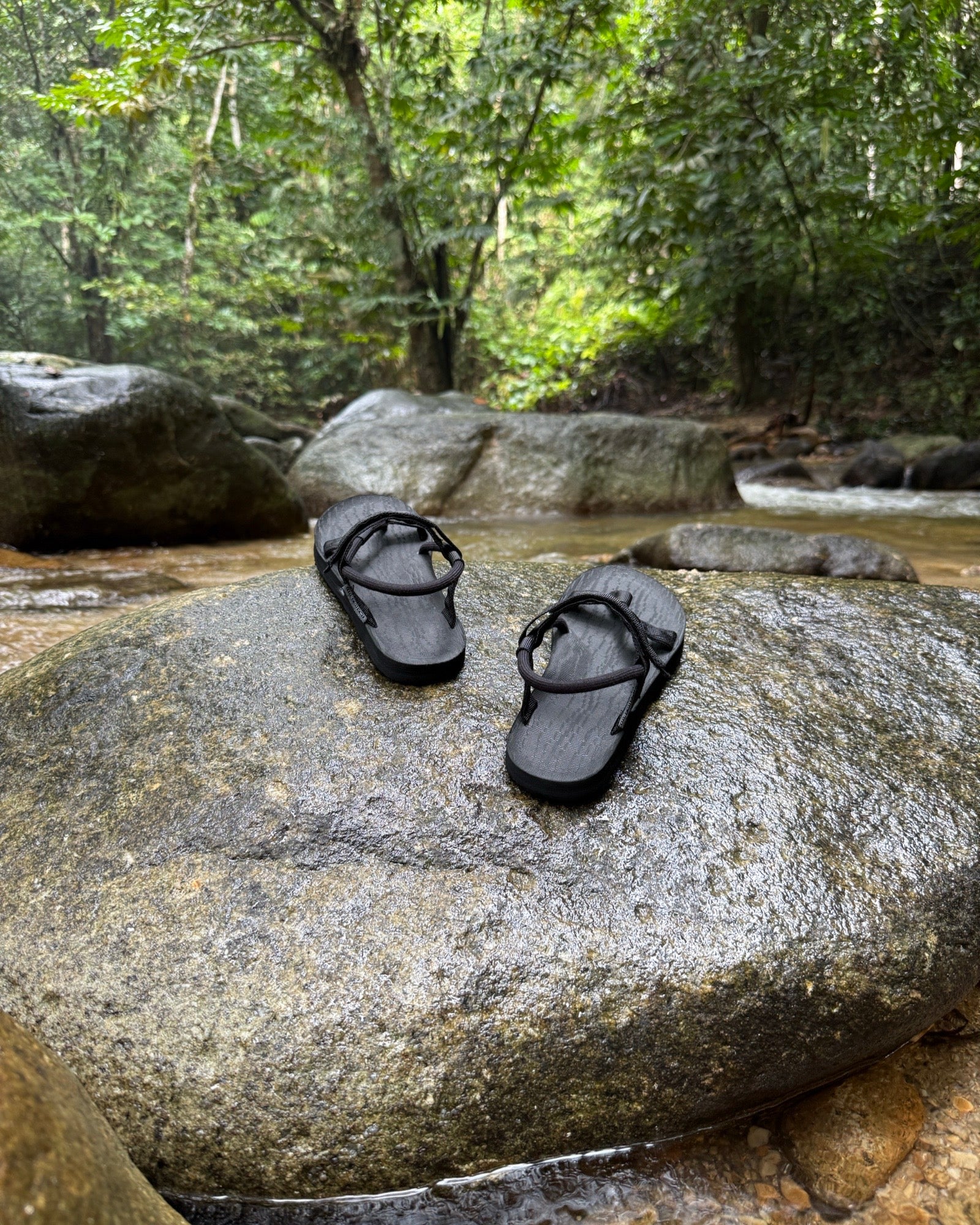 Women's matt black EXPLOre Roman Sandals on a river rock with a lush tropical rainforest backdrop, the perfect footwear for authentic tropical exploration.