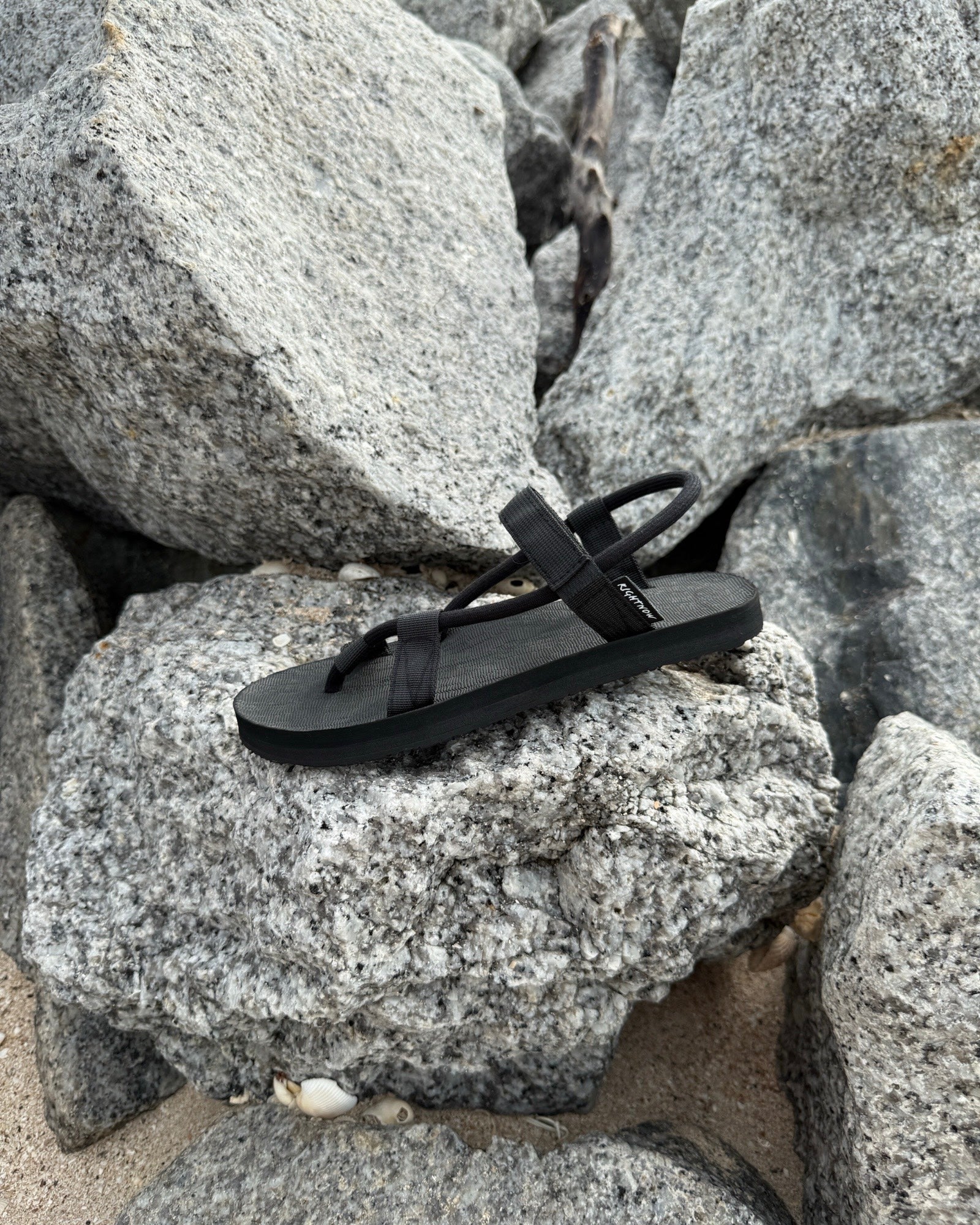 A single women's matt black EXPLOre Roman Sandal on a rugged coastal granite boulder, highlighting its durable minimalist strap design for all-terrain wear.