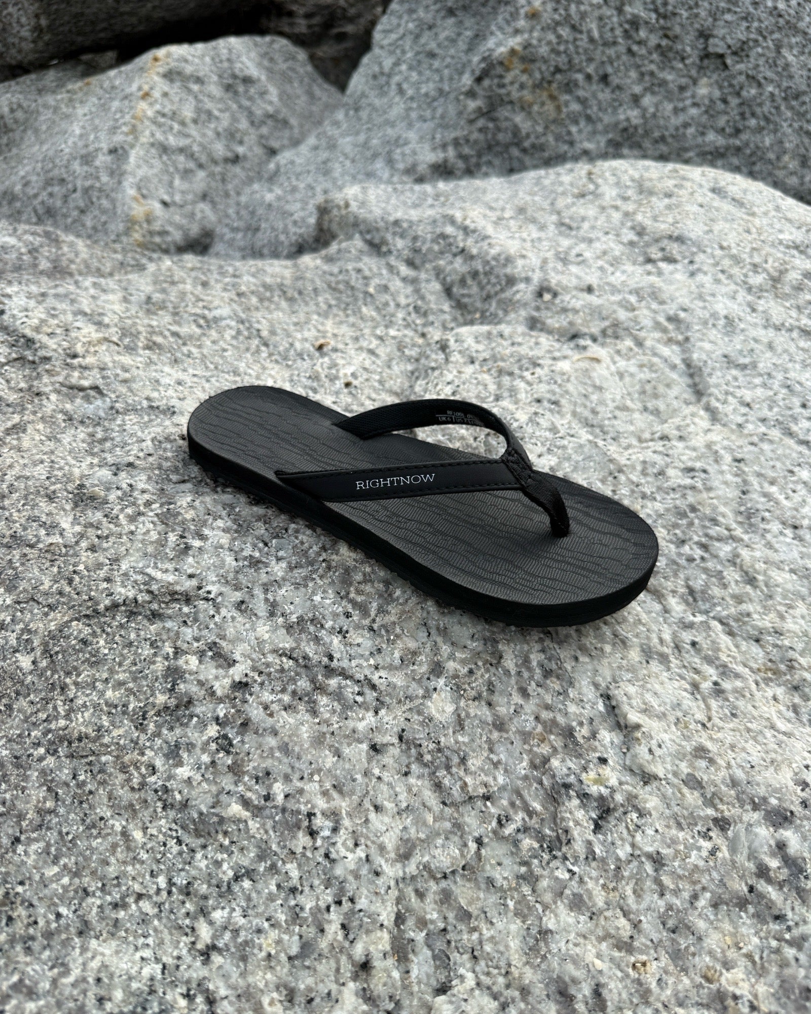 A single women's matt black Flow Thong Sandal resting on rugged coastal rock, showcasing its minimalist design for effortless outdoor presence.