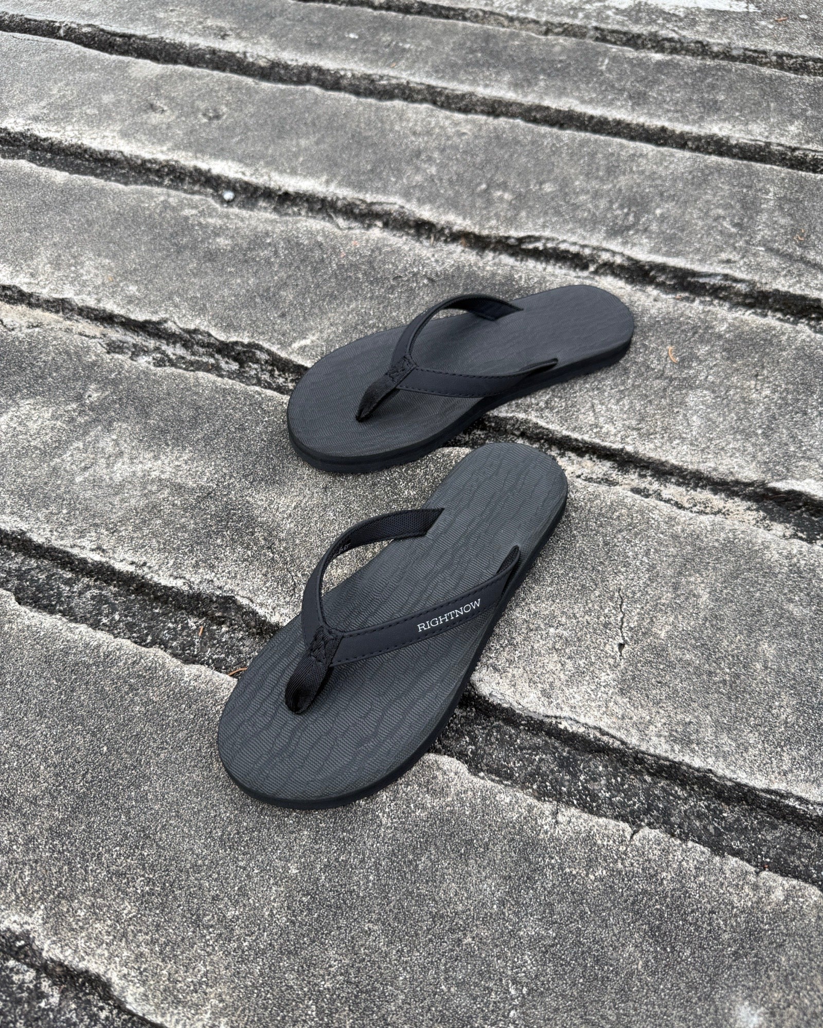 A pair of women's matt black Flow Thong Sandals on a dark, textured concrete surface, showcasing their minimalist comfort for modern living.