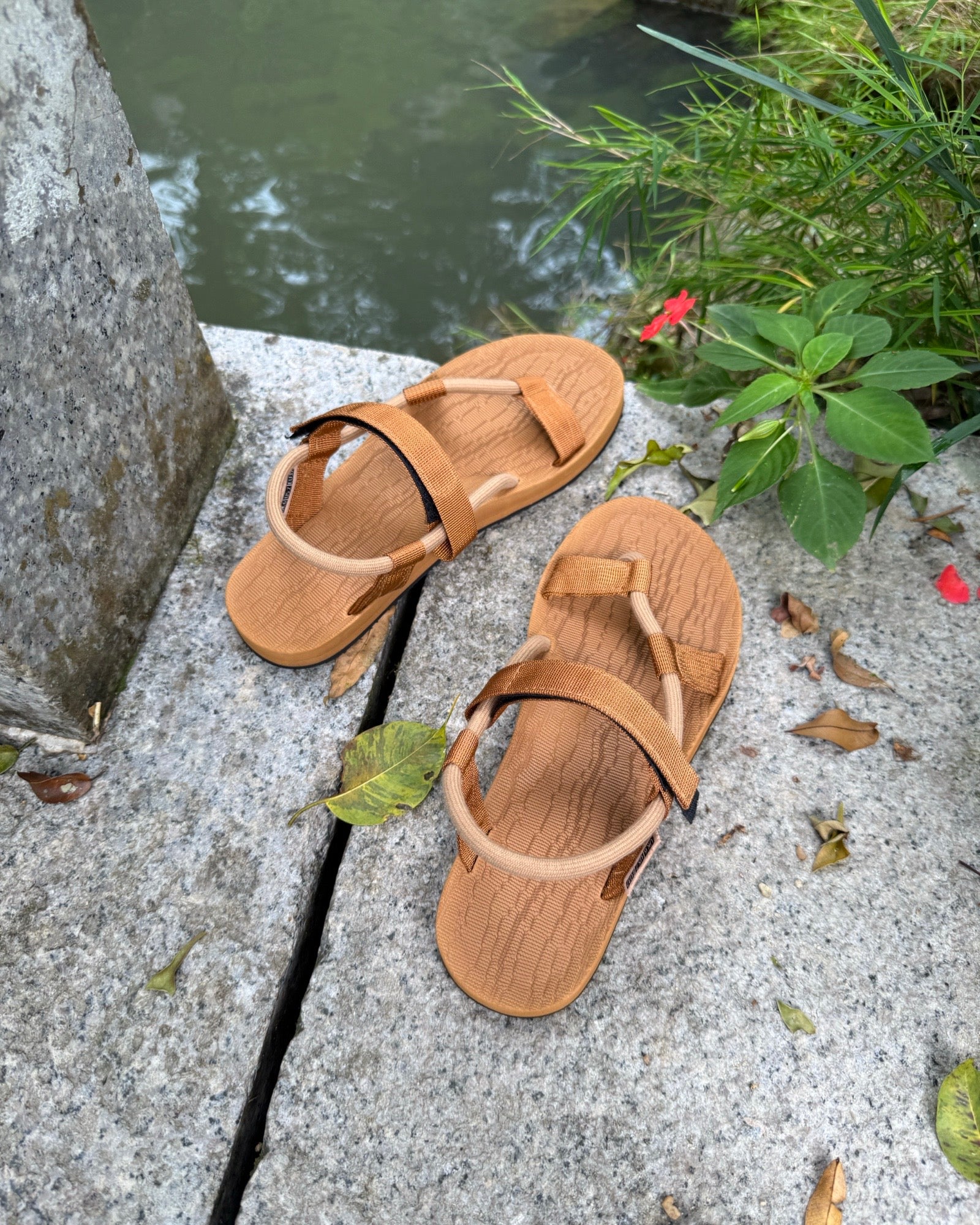 A pair of women's earth brown EXPLOre Roman Sandals on a stone ledge by a tranquil stream, showing a simple, authentic lifestyle connected with nature.