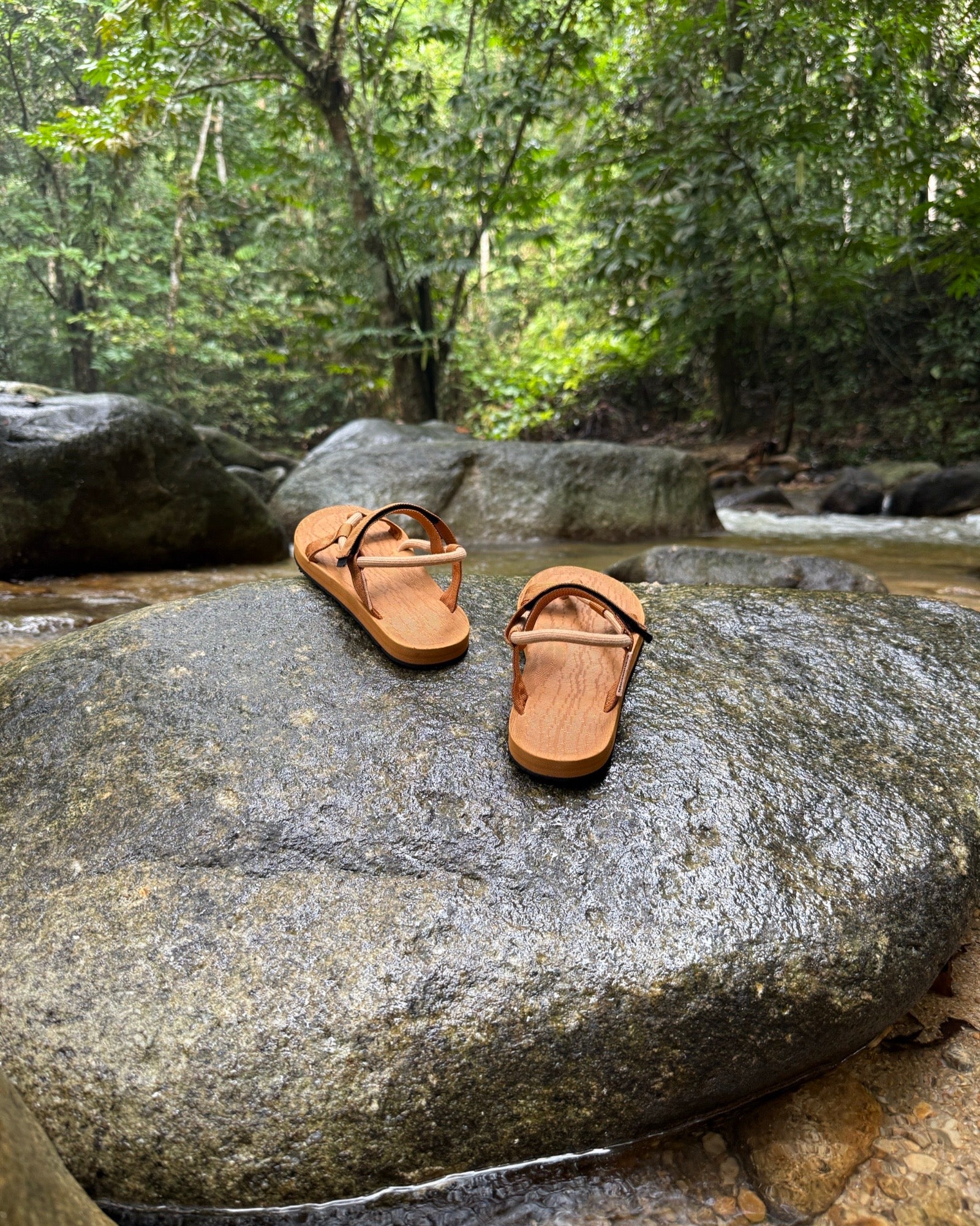 Women's earth brown EXPLOre Roman Sandals on a river rock with a lush tropical rainforest backdrop, the perfect footwear for authentic tropical exploration.
