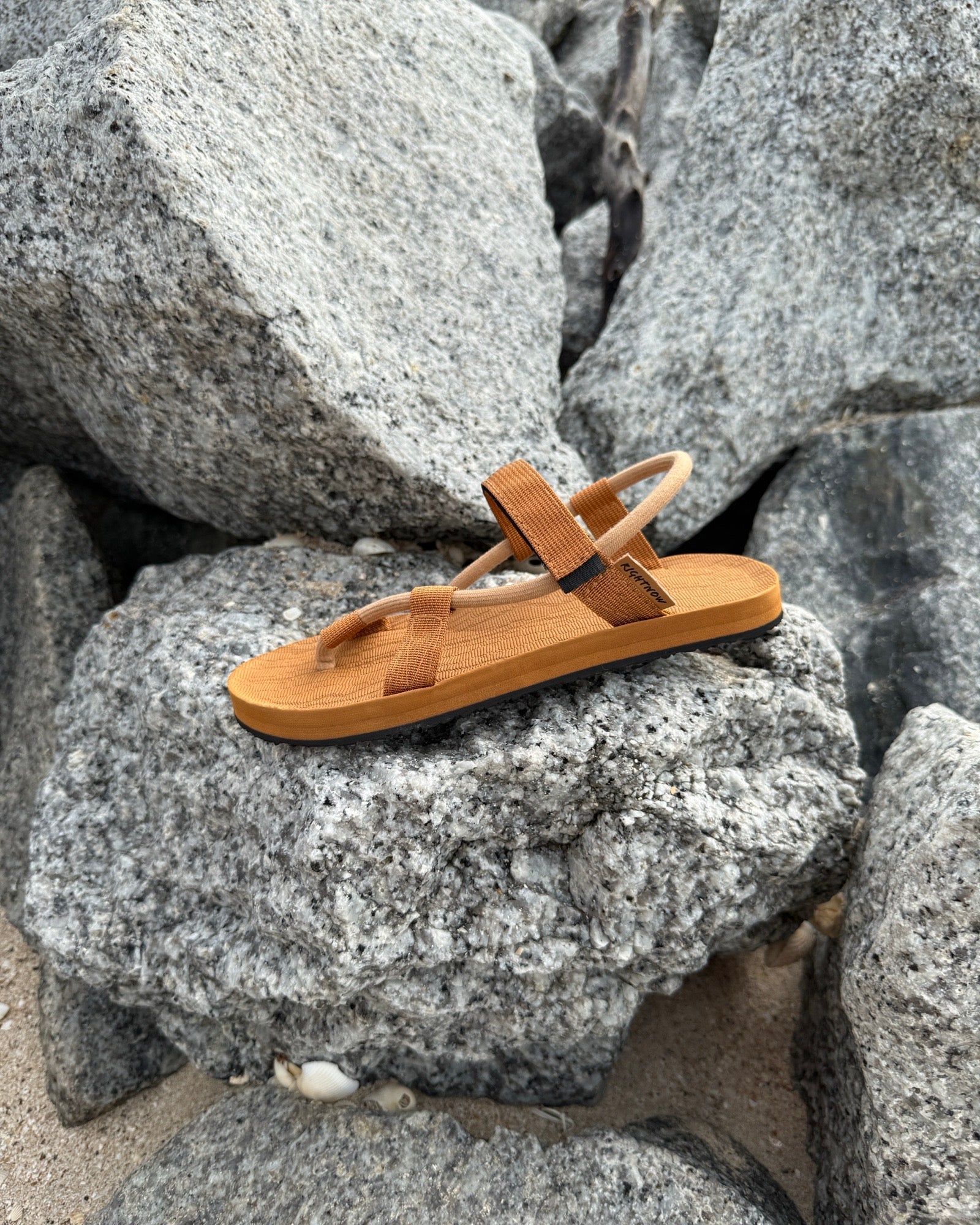 A single women's earth brown EXPLOre Roman Sandal on a rugged coastal granite boulder, highlighting its durable minimalist strap design for all-terrain wear.