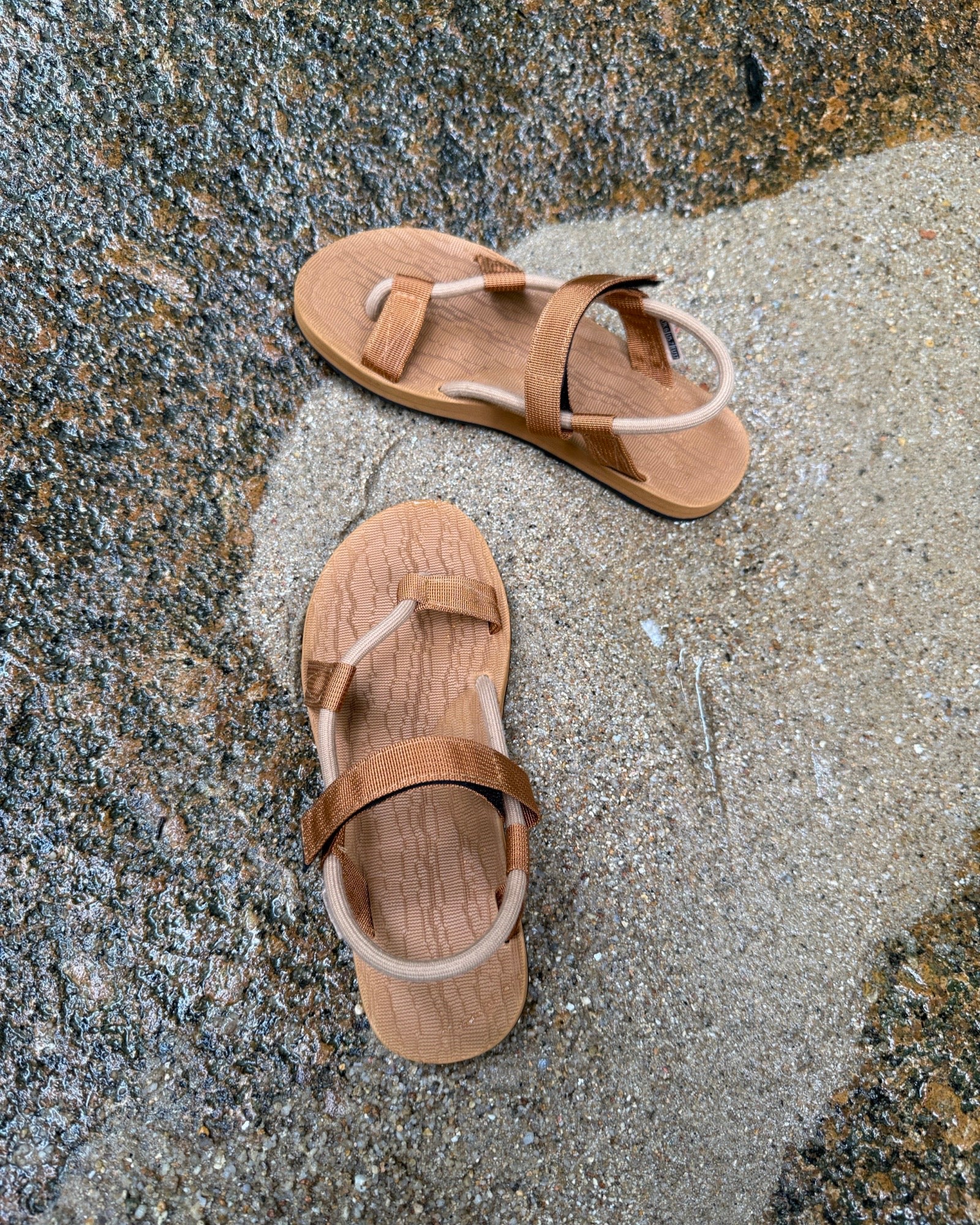Top-down view of women's earth-tone EXPLOre Roman Sandals on wet, sandy riverside ground, showing their simple design and flexible straps for any natural terrain.