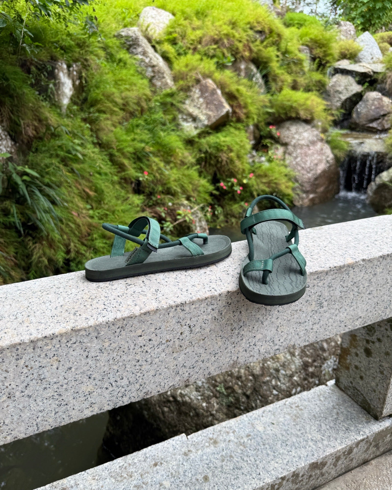 Women's natural green EXPLOre Roman Sandals on a bridge over a serene waterfall and lush greenery, perfect for travel and finding a quiet moment of presence in nature.