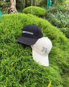 The black RIGHTNOW logo cap and the beige RN monogram cap shown together on a vibrant green grassy mound, representing simple, authentic style for everyone.