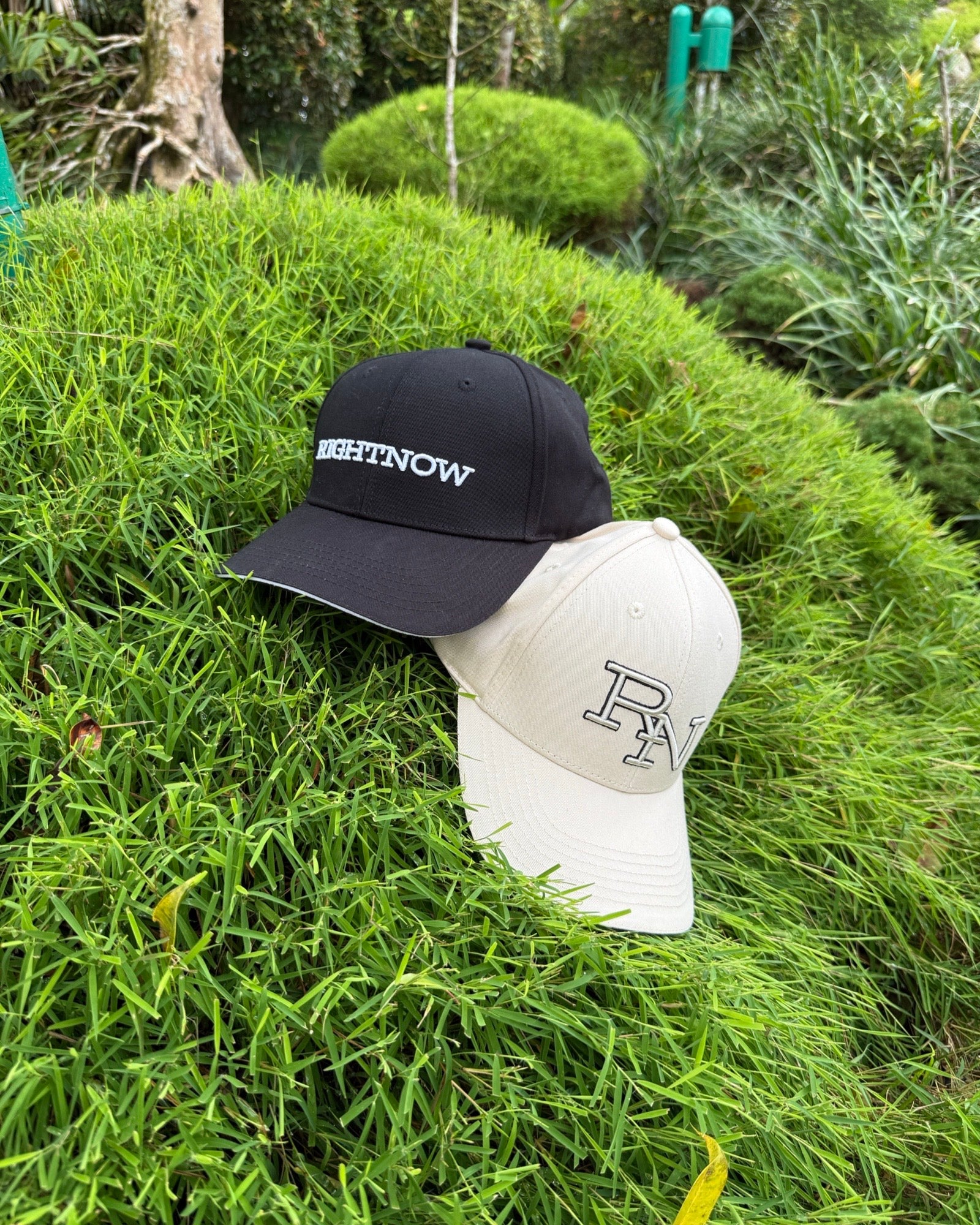 The black RIGHTNOW logo cap and the beige RN monogram cap shown together on a vibrant green grassy mound, representing simple, authentic style for everyone.