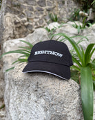 A matt black Ignite RIGHTNOW logo cap resting on a large, sunlit rock in a quiet garden, embodying a moment of simple, peaceful presence outdoors.
