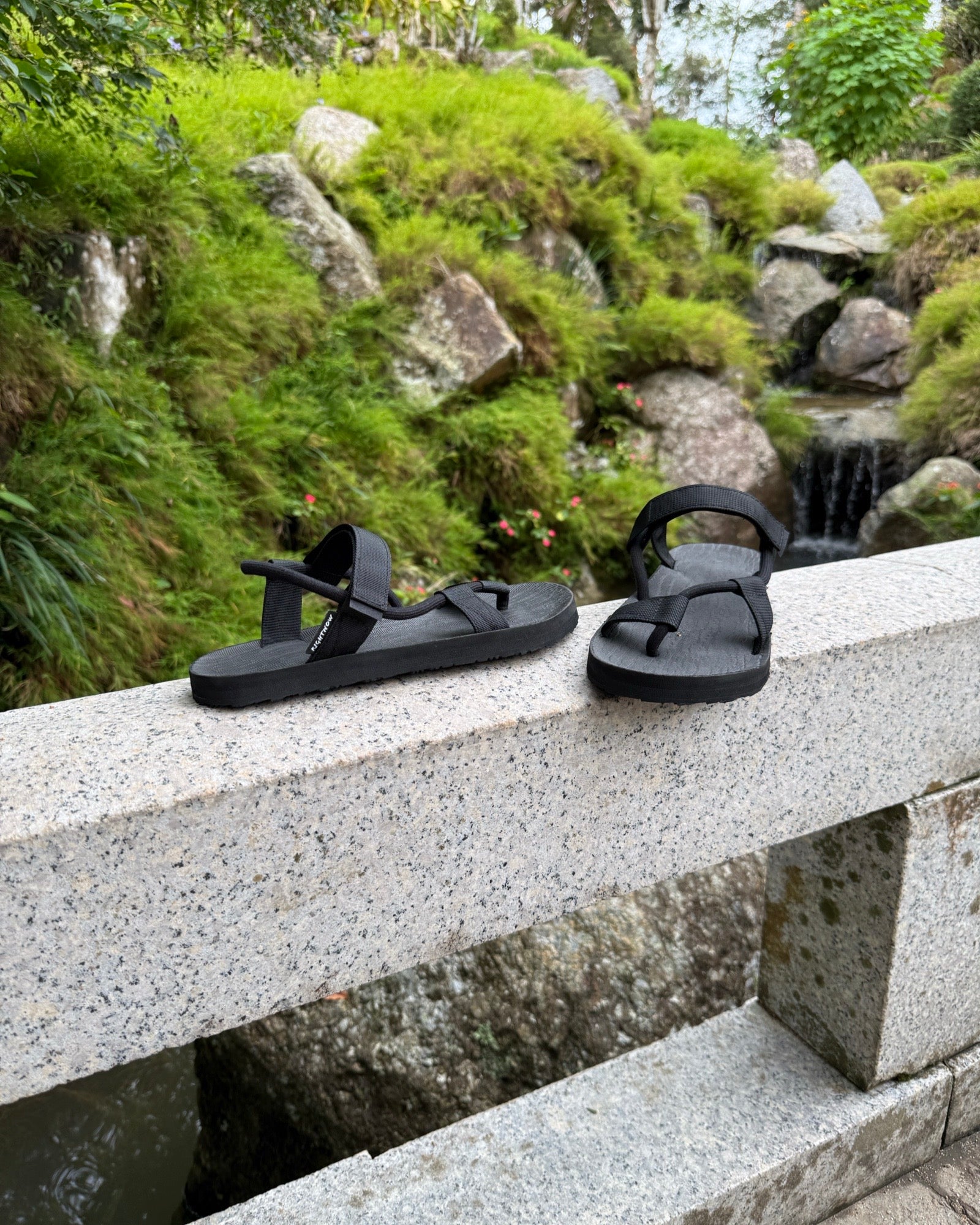 Men's matt black EXPLOre Roman Sandals on a bridge over a serene waterfall and lush greenery, perfect for travel and finding a quiet moment of presence in nature.