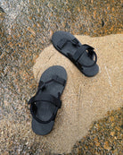 Top-down view of men's elegant-tone EXPLOre Roman Sandals on wet, sandy riverside ground, showing their simple design and flexible straps for any natural terrain.