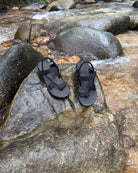 Waterproof men's matt black EXPLOre Roman Sandals on a wet boulder in a flowing jungle stream, highlighting their anti-slip soles for secure footing on any terrain.