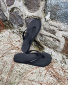 Men's matt black Flow Thong Sandals leaning against a textured stone wall, conveying a casual yet authentic style ready for everyday adventures.