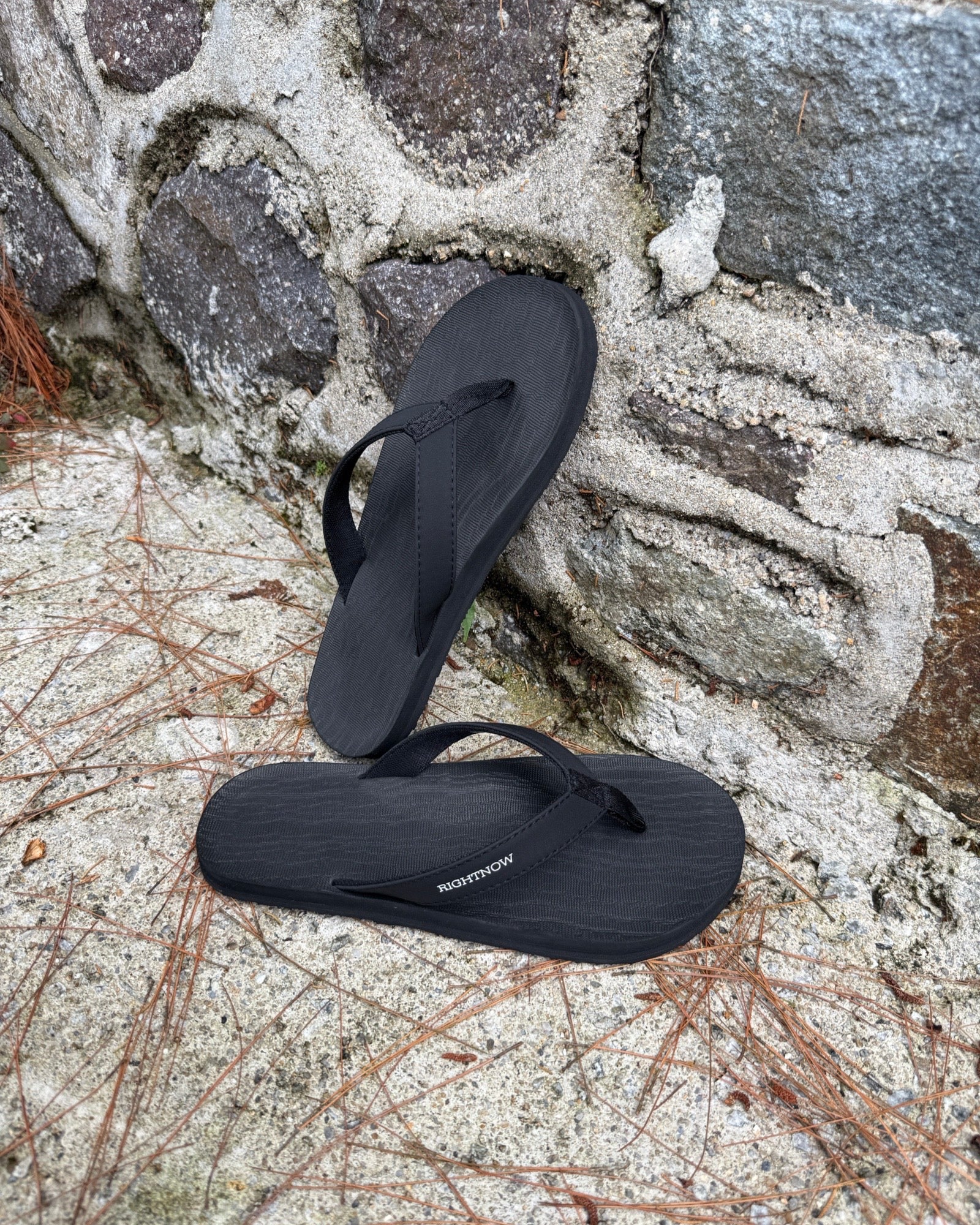 Men's matt black Flow Thong Sandals leaning against a textured stone wall, conveying a casual yet authentic style ready for everyday adventures.