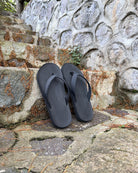 A pair of men's matt black Flow Thong Sandals on rustic stone steps, perfect for a simple and authentic journey through historical or natural paths.