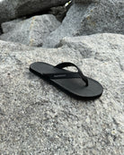 A single men's matt black Flow Thong Sandal resting on rugged coastal rock, showcasing its minimalist design for effortless outdoor presence.