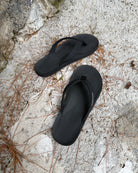 Top-down view of men's elegant-tone Flow Thong Sandals on concrete ground with scattered pine needles, highlighting their simple design for natural and urban exploration.