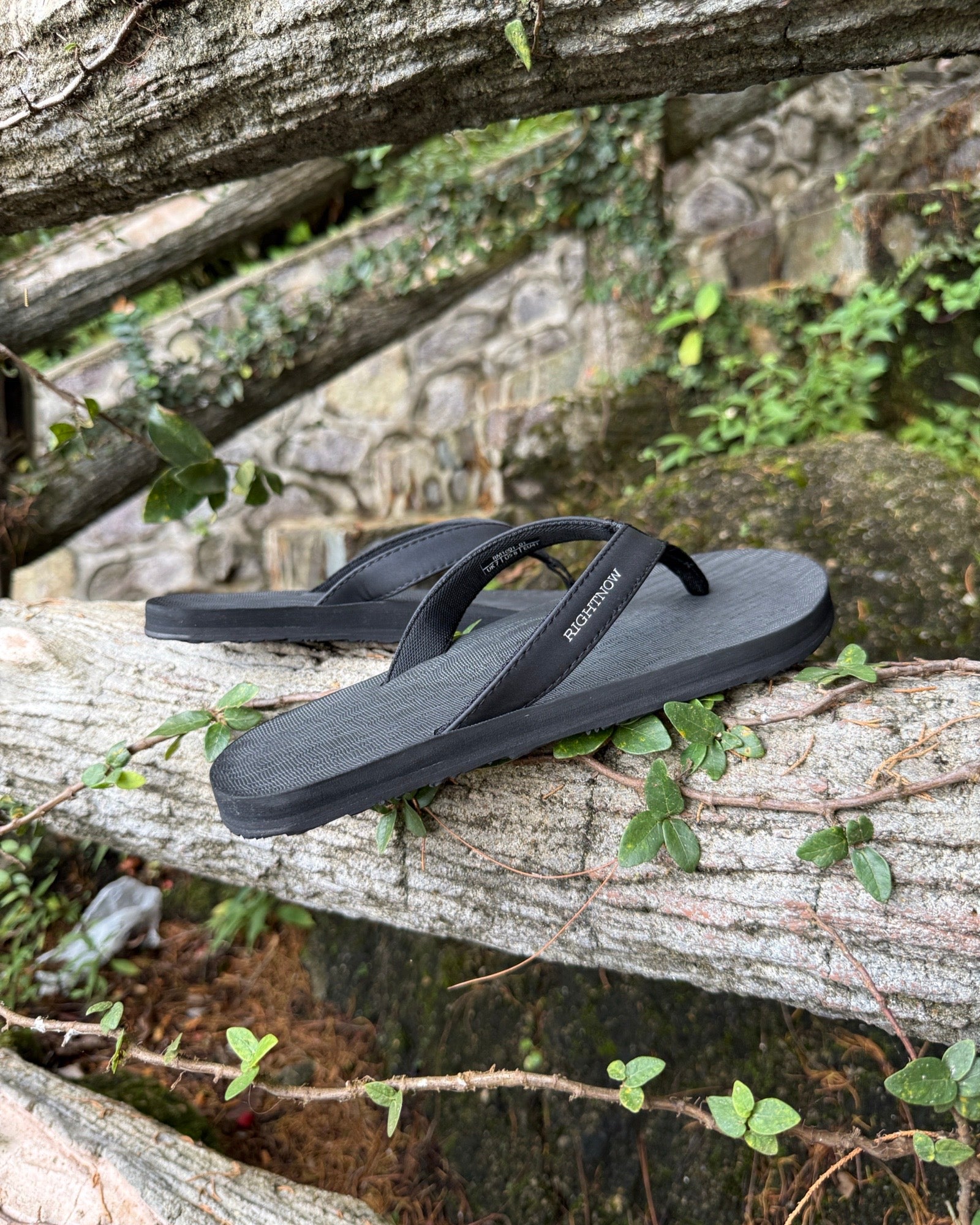 Men's matt black Flow Thong Sandals displayed on a moss-covered fallen log amidst lush greenery, perfect for authentic moments of relaxation at a campground in nature.
