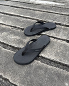 A pair of men's matt black Flow Thong Sandals on a dark, textured concrete surface, showcasing their minimalist comfort for modern living.