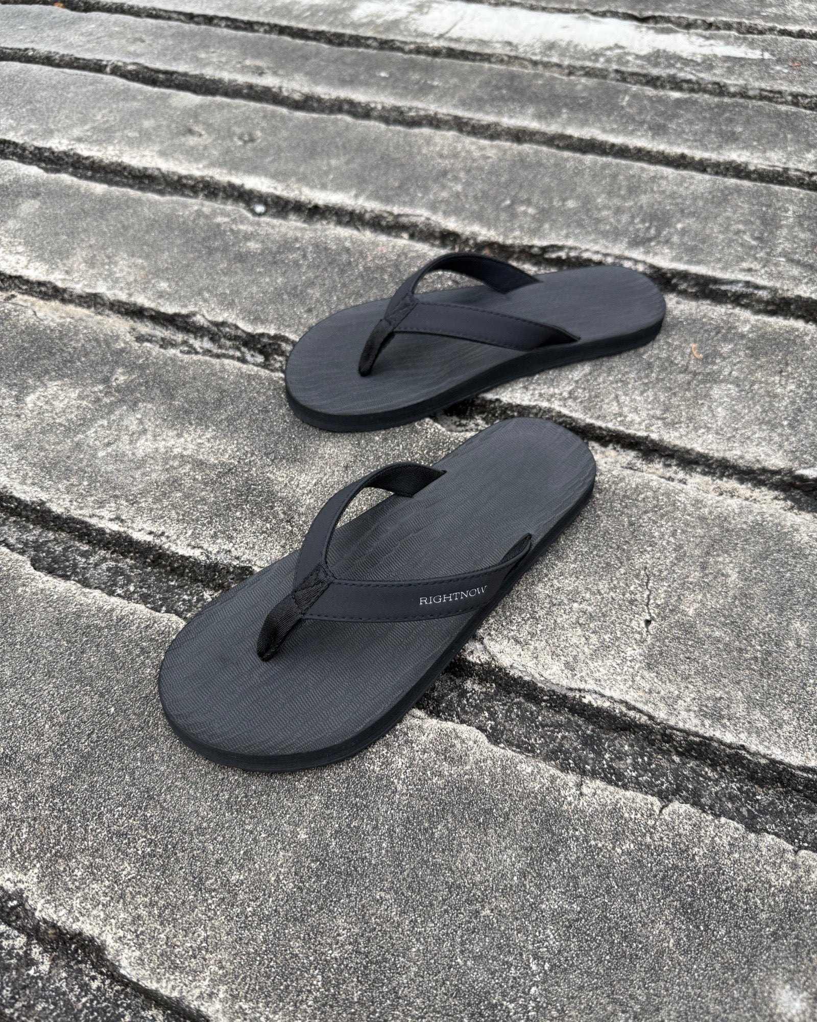 A pair of men's matt black Flow Thong Sandals on a dark, textured concrete surface, showcasing their minimalist comfort for modern living.