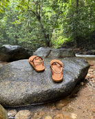 Men's earth brown EXPLOre Roman Sandals on a river rock with a lush tropical rainforest backdrop, the perfect footwear for authentic tropical exploration.