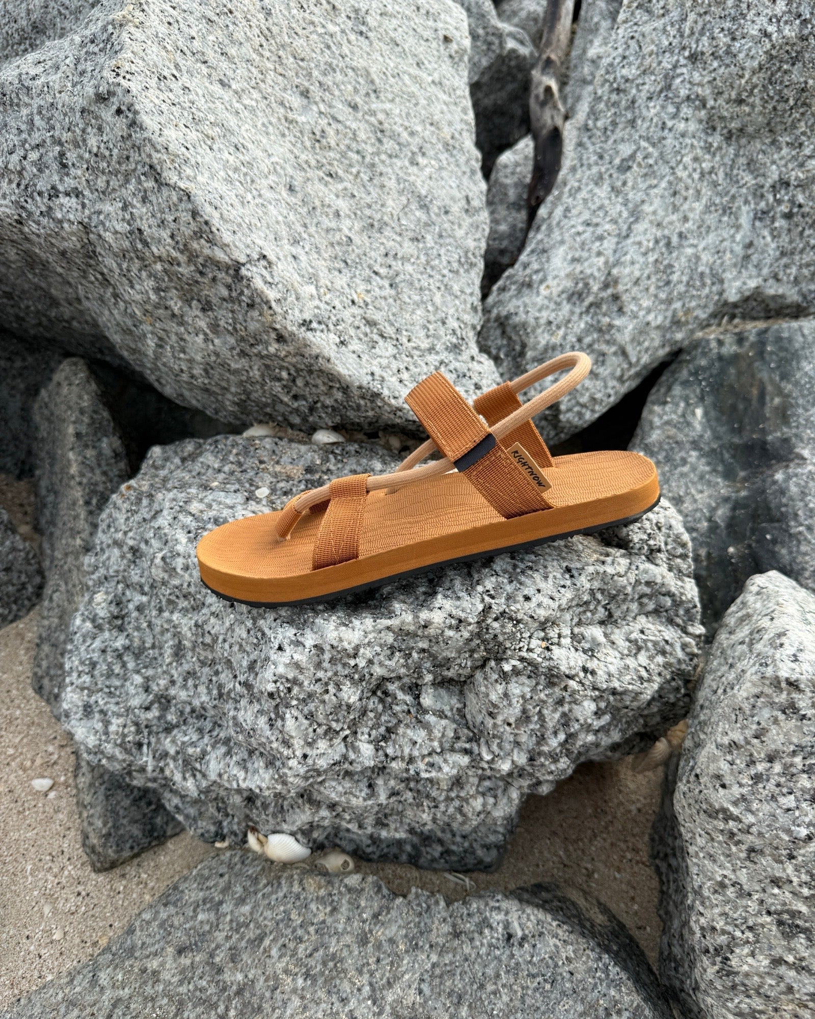 A single men's earth brown EXPLOre Roman Sandal on a rugged coastal granite boulder, highlighting its durable minimalist strap design for all-terrain wear.