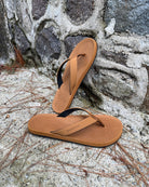 Men's earth brown Flow Thong Sandals leaning against a textured stone wall, conveying a casual yet authentic style ready for everyday adventures.
