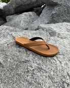 A single men's earth brown Flow Thong Sandal resting on rugged coastal rock, showcasing its minimalist design for effortless outdoor presence.