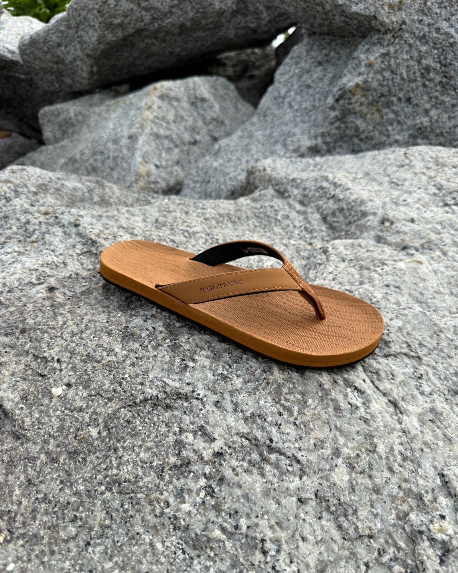 A single men's earth brown Flow Thong Sandal resting on rugged coastal rock, showcasing its minimalist design for effortless outdoor presence.