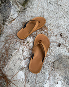 Top-down view of men's earth-tone Flow Thong Sandals on concrete ground with scattered pine needles, highlighting their simple design for natural and urban exploration.