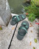 A pair of men's natural green EXPLOre Roman Sandals on a stone ledge by a tranquil stream, showing a simple, authentic lifestyle connected with nature.