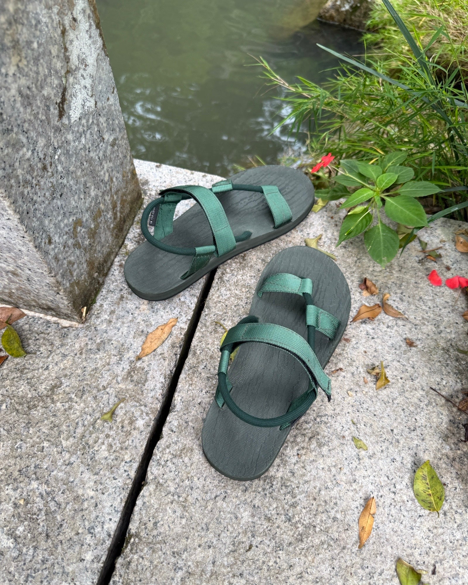A pair of men's natural green EXPLOre Roman Sandals on a stone ledge by a tranquil stream, showing a simple, authentic lifestyle connected with nature.