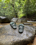 Men's natural green EXPLOre Roman Sandals on a river rock with a lush tropical rainforest backdrop, the perfect footwear for authentic tropical exploration.