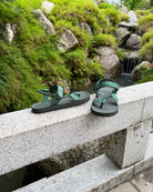 Men's natural green EXPLOre Roman Sandals on a bridge over a serene waterfall and lush greenery, perfect for travel and finding a quiet moment of presence in nature.